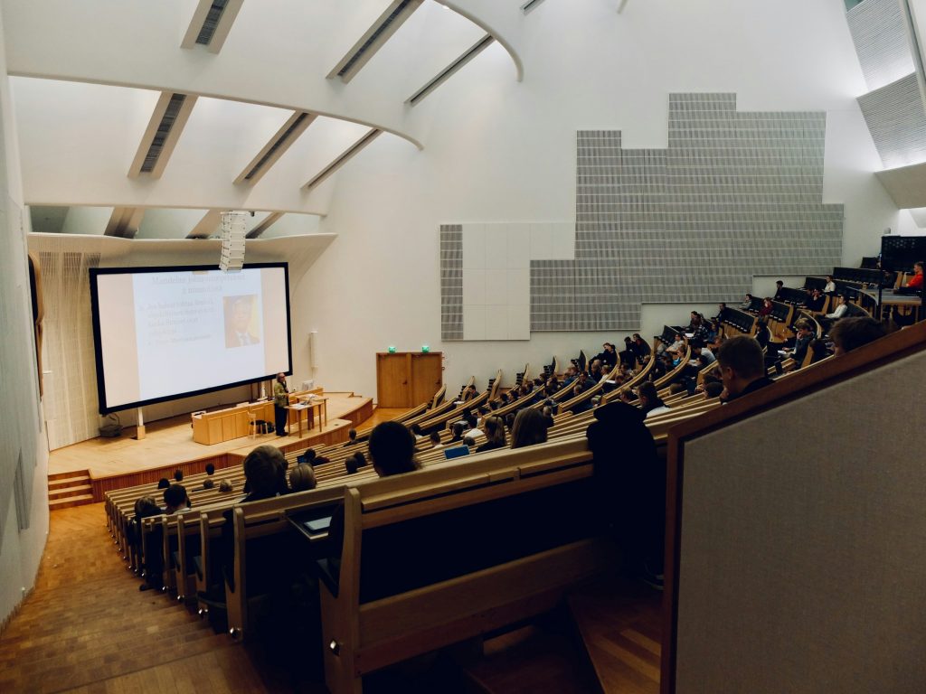 University students attending a lecture in a modern auditorium with tiered seating and a large presentation screen.