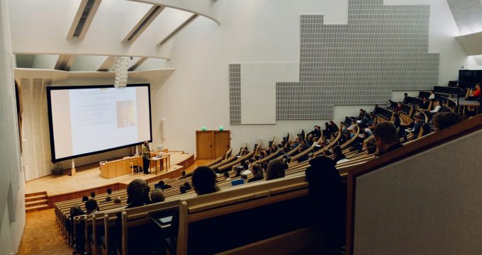 University students attending a lecture in a modern auditorium with tiered seating and a large presentation screen.